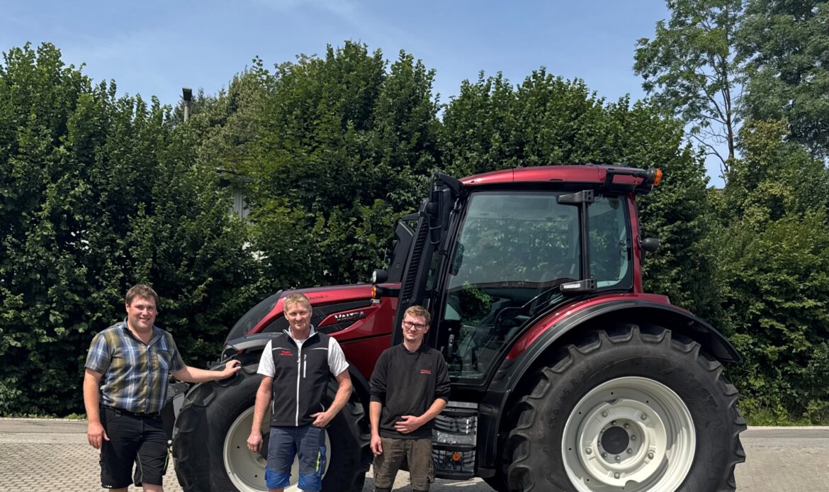 Foto einer Maschinenübergabe. Man sieht unseren Verkäufer Martin mit dem Kunden Rainer Michael + Begleitung vor einem roten Valtra Schlepper N155A.