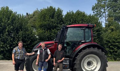 Foto einer Maschinenübergabe. Man sieht unseren Verkäufer Martin mit dem Kunden Rainer Michael + Begleitung vor einem roten Valtra Schlepper N155A.