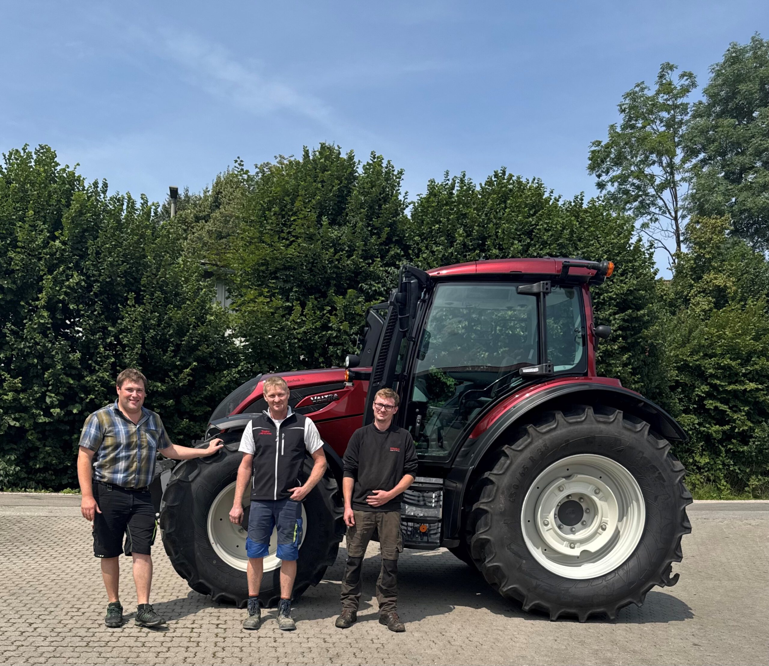 Foto einer Maschinenübergabe. Man sieht unseren Verkäufer Martin mit dem Kunden Rainer Michael + Begleitung vor einem roten Valtra Schlepper N155A.